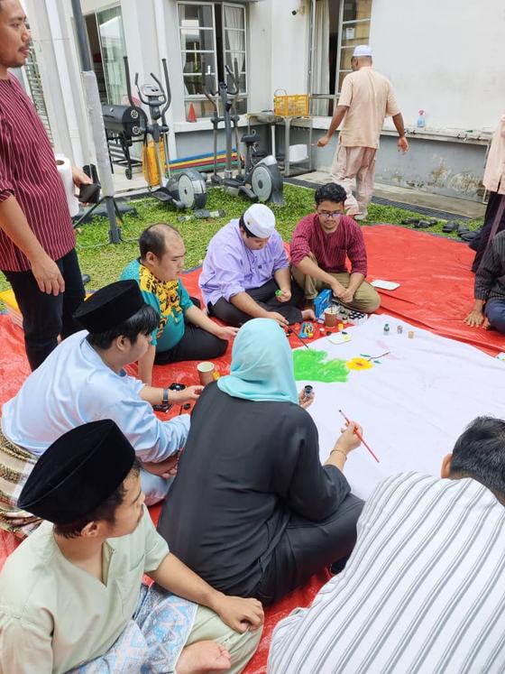 Pelatih, tenaga pengajar dan ibu bapa duduk berkumpulan melukis sepanduk kain di atas hamparan.